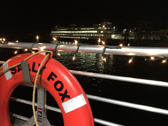 The railing of the Sally Fox is decked in holiday lights, with a Washington State Ferry in the background.